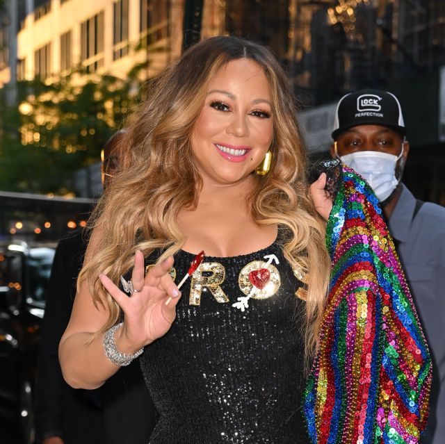 mariah carey smiles and waves at the camera while walking on a city sidewalk, she holds a multicolored sequin coat over one shoulder and wears a black sequin sleevless minidress