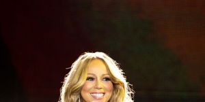 mariah carey smiles and holds a microphone to her face with her other hand on her hip, she wears a black strapless top with a silver sequin skirt