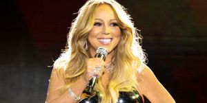mariah carey smiles and holds a microphone to her face with her other hand on her hip, she wears a black strapless top with a silver sequin skirt