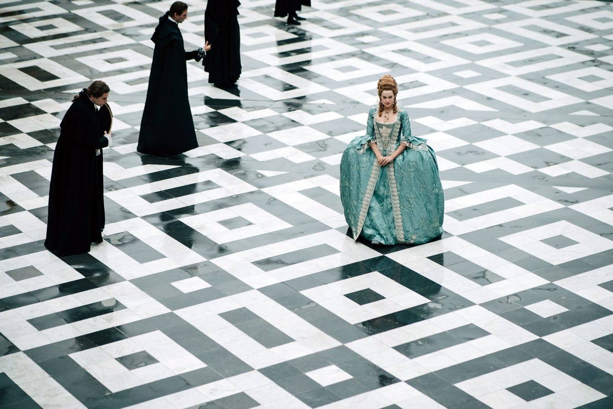a publicity photo of emilia schule as marie antoinette, on a black and white tiled floor, with four men in black standing behind her