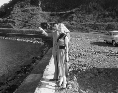 original caption 2101954 japan baseball player joe dimaggio and his wife actress and sex symbol marilyn monroe are shown visiting a little fishing village standing by a body of water a small hill and trees are visible in the background as dimaggio points out a sight to monroe inp photograph