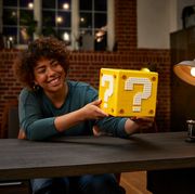 a woman smiles and displays the lego super mario 64 question mark cube, sitting at a desk in an open plan apartment