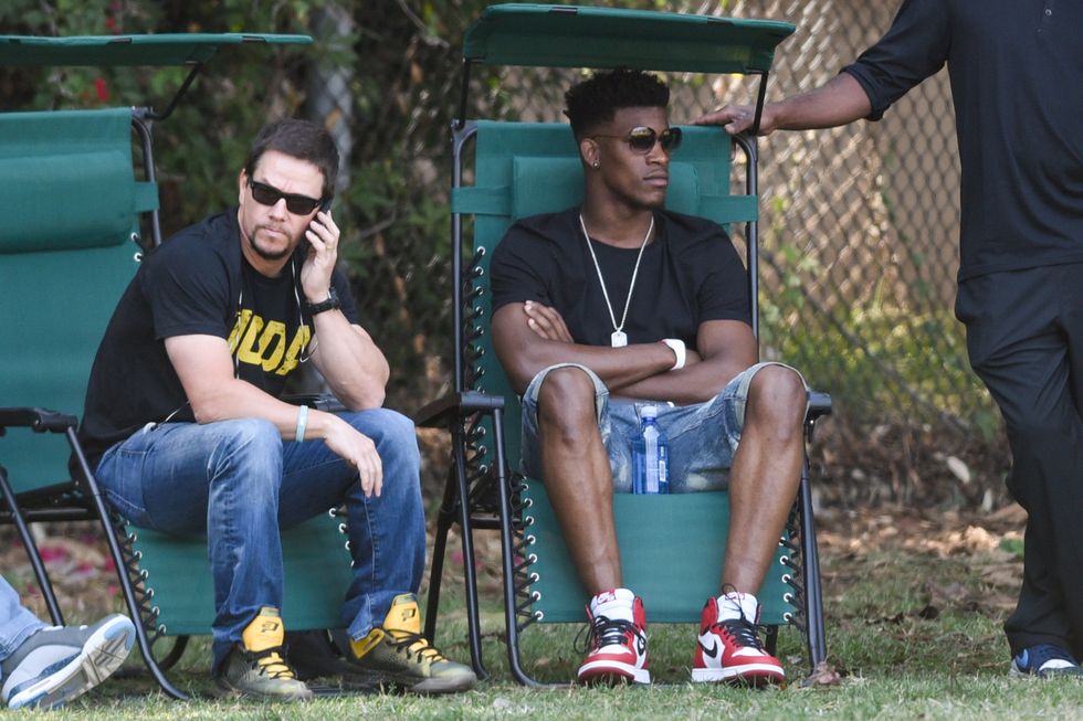 mark wahlberg and jimmy butler, both wearing black t shirts, jeans, and sunglasses, sit on green lawn chairs outside on the grass