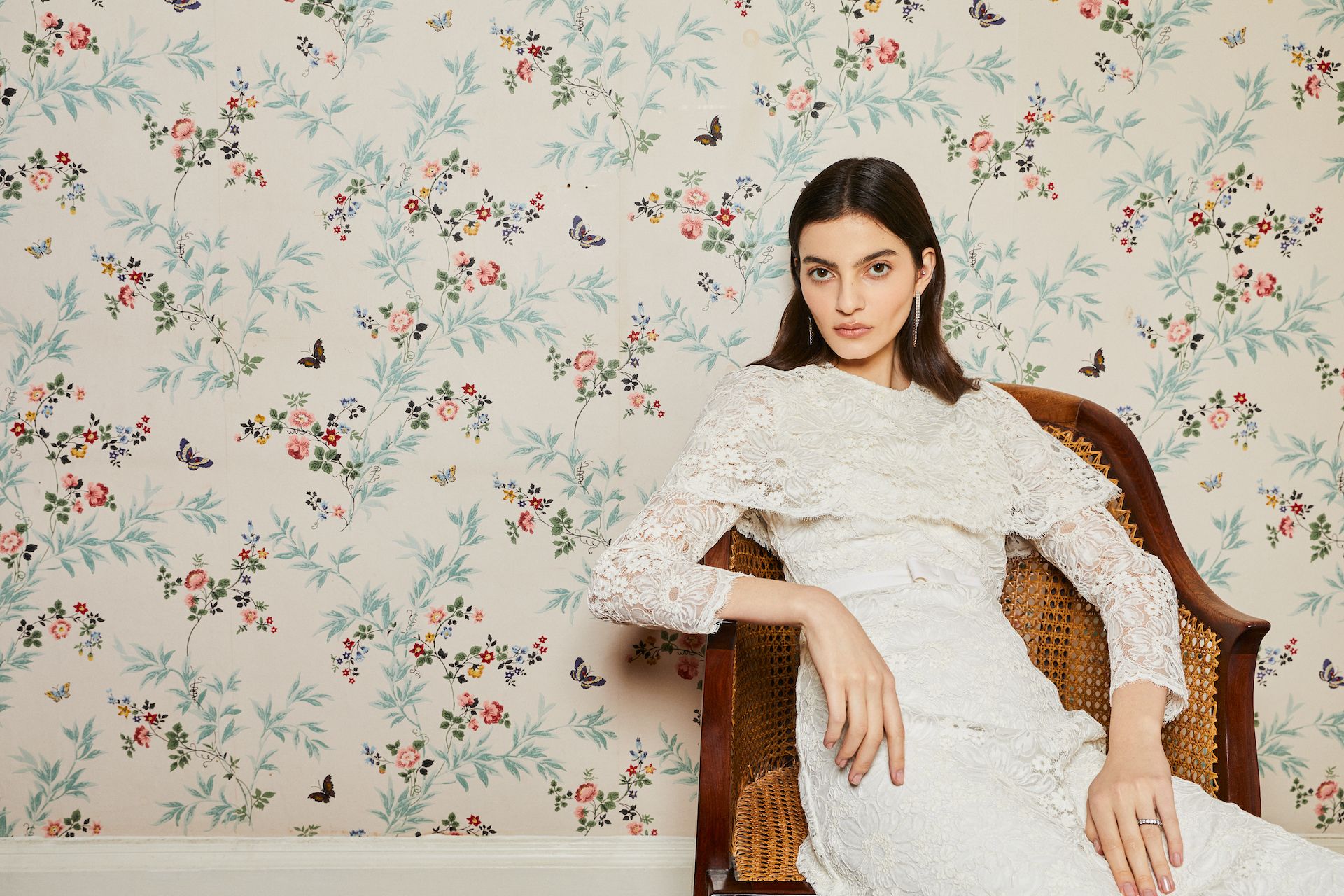 a brunette model wears a lace dress in front of a floral wallpaper background