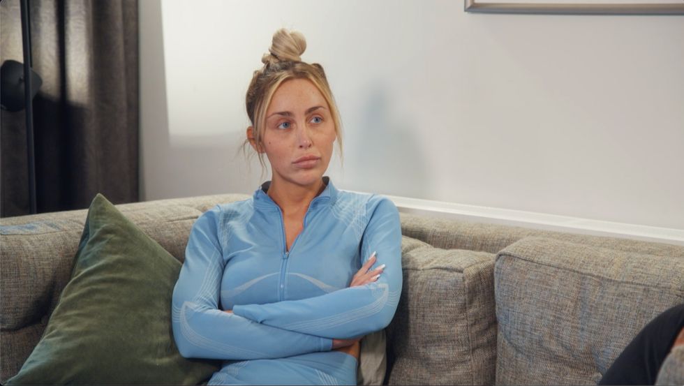 ella aus „Married at First Sight UK“, Staffel 8, Folge 18, ist eine Frau mit blauen Augen und blonden Haaren, die zu einem Haarknoten hochgesteckt sind. Sie trägt ein blaues Trainingsoberteil und sitzt mit vor der Brust verschränkten Armen auf einer grauen Couch