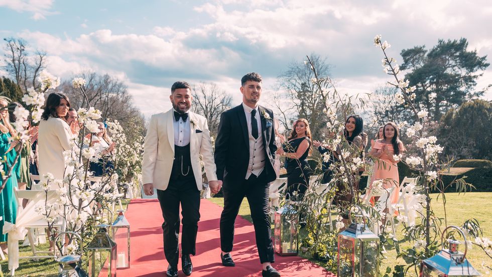 Mark und Sean, zwei Männer in Anzügen, die über einen roten Teppich laufen, umgeben von Blumen und Gästen auf einem Feld, bei ihrer Hochzeit, ausgestrahlt auf „Married At First Sight UK“, Staffel 8, Folge 18