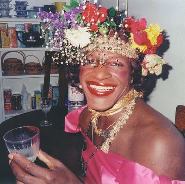 marsha p johnson smiles at the camera while seated at a table, she wears a flower crown and a bright pink dress and holds a glass