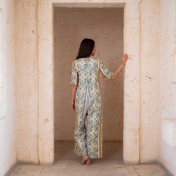 a figure stands with her back facing the viewer in a spacious, light colored corridor framed by stone walls