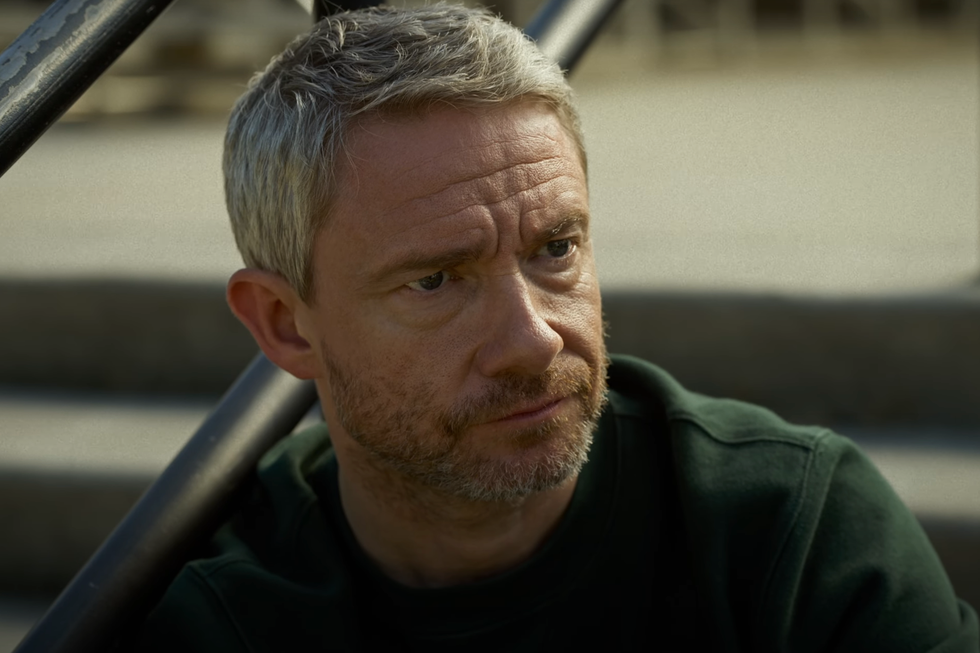 Martin Freeman, ein Mann mit grauen Haaren und einem dunkelgrünen Pullover, sitzt auf Betonstufen und grinst in einer Szene aus „Miller's Girl“ jemanden außerhalb der Kamera an