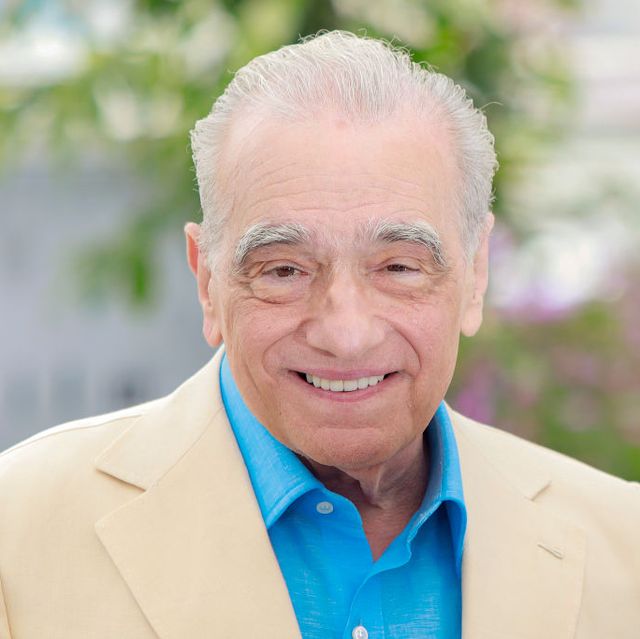 martin scorsese smiles at the camera, he wears a cream suit jacket with a bright blue collared shirt unbuttoned at the neck