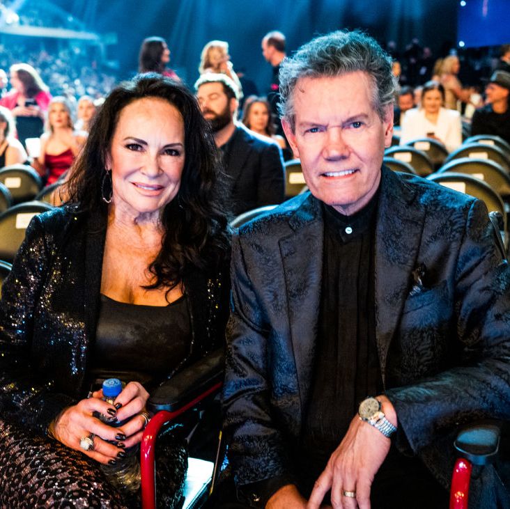 mary davis and randy travis smile at the camera while seated, he wears a black shirt and dark navy patterned suit jacket with a silver watch, she wears a black shirt, a black sequin jacket, and black and silver patterned pants, behind them are chairs and additional people seated
