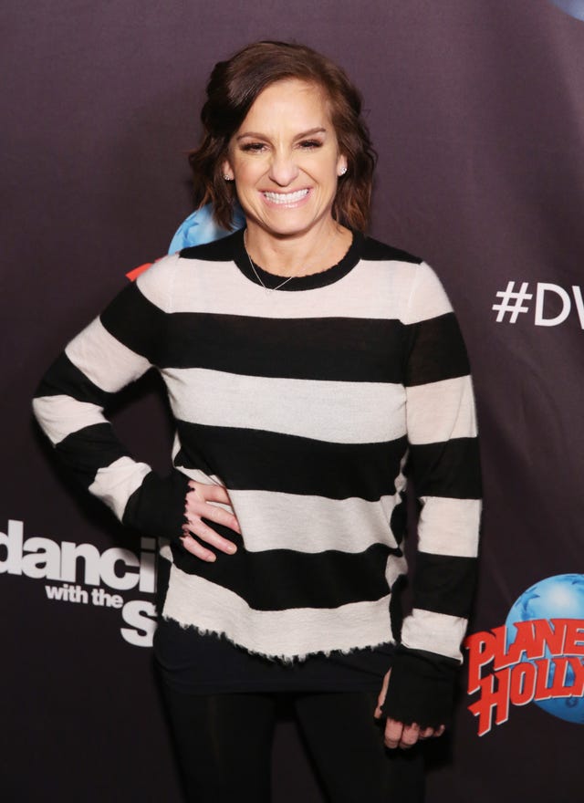 mary lou retton smiles and stands with one hand on her hip, she wears a black and cream colored shirt and small silver necklace