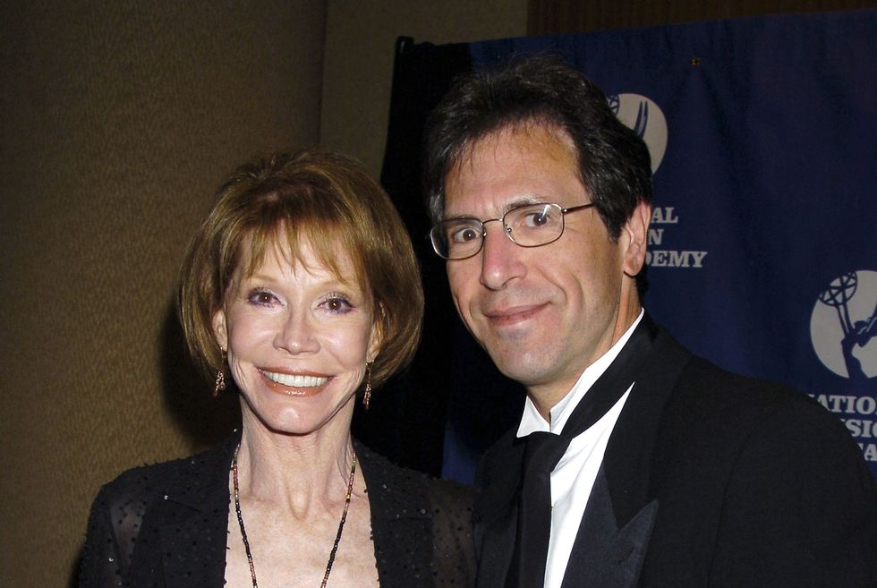 mary tyler moore and husband richard levine smile at the camera, she wears a black sweater over a blush top with jewelry, he wears a black suit with a white shirt