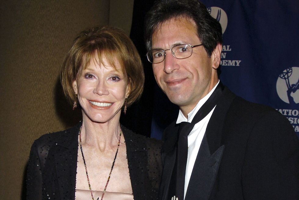 mary tyler moore and husband richard levine smile at the camera, she wears a black sweater over a blush top with jewelry, he wears a black suit with a white shirt
