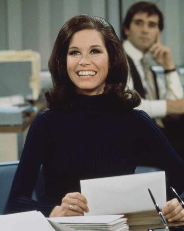 mary tyler moore on set of the mary tyler moore show, she sits at a desk and holds an envelope as she looks upward and smiles