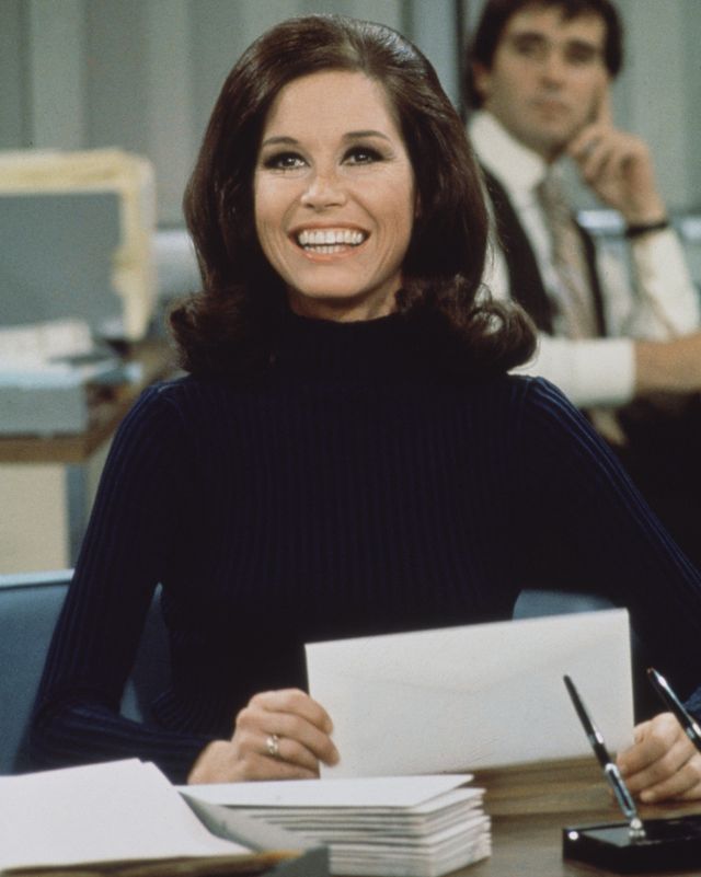 mary tyler moore on set of the mary tyler moore show, she sits at a desk and holds an envelope as she looks upward and smiles