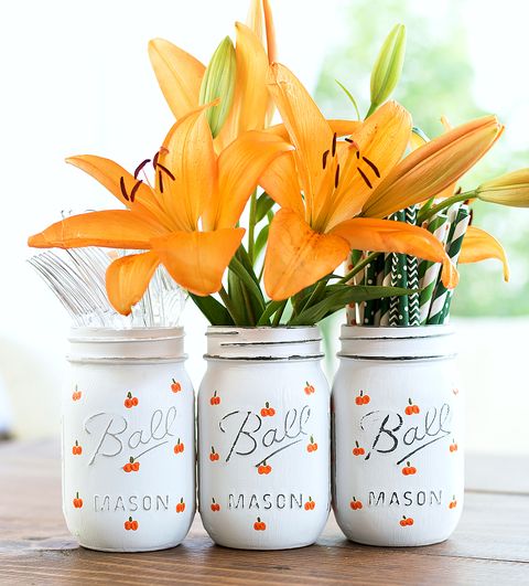 three mason jars in a row painted white and with small orange pumpkins, one is holding clear forks, one orange flowers and one straws