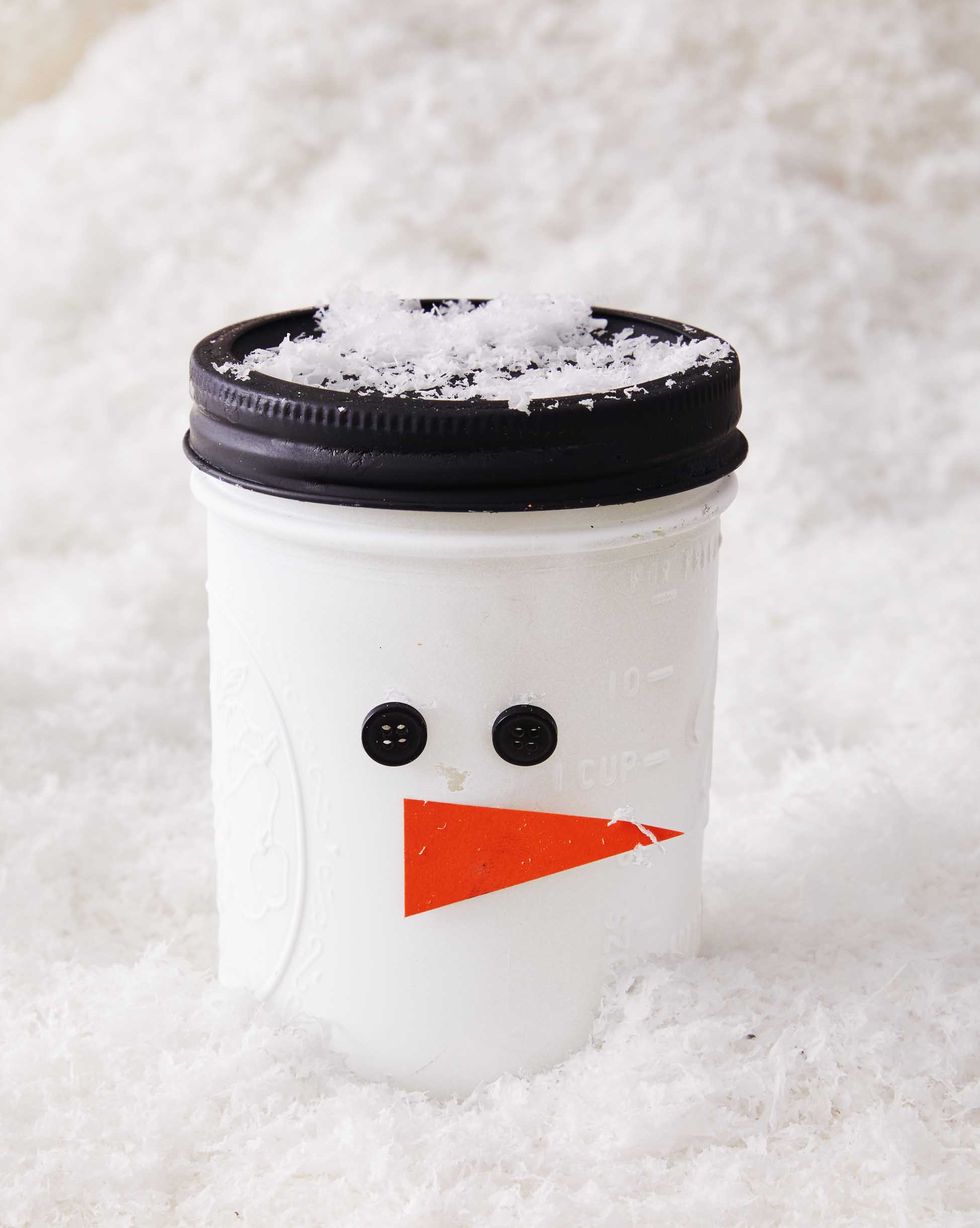 a mason jar painted white with a black painted lid with button eyes and a paper nose meant to look like a snowman
