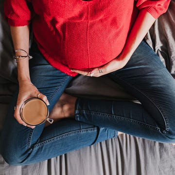 top view of pregnant woman in red sweater holding their baby bump while sitting cross legged in jeans on a bed and holding a mug of coffee