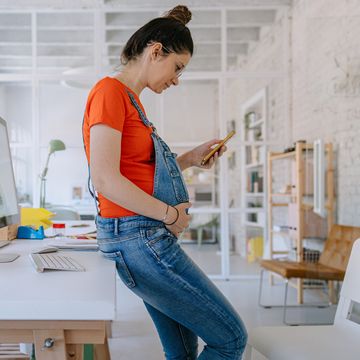 pregnant person in maternity overalls looking at her phone in a workspace