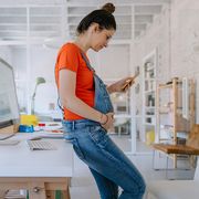 pregnant person in maternity overalls looking at her phone in a workspace