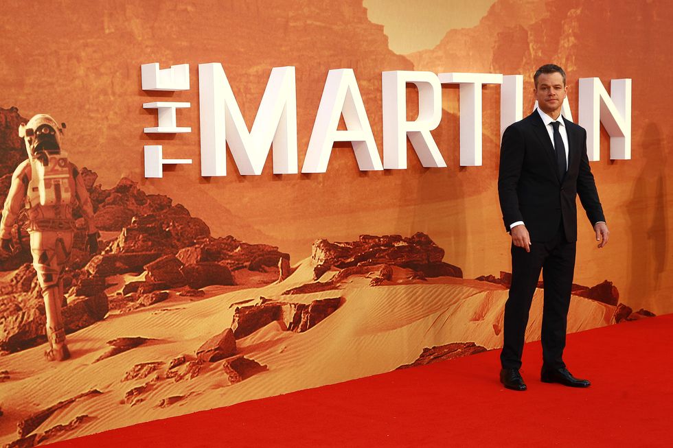 matt damon, wearing a black suit and tie, standing in front of a large promotional display for the film the martian