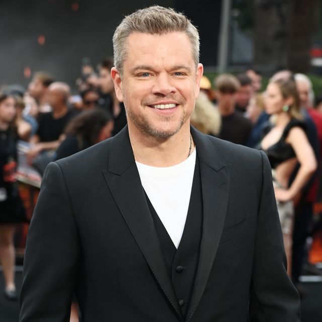 matt damon smiles at the camera, he wears a black suit jacket over a black vest and white shirt, behind him is a blurred out crowd of people