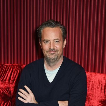 matthew perry sits with his arms folded against a red sofa with a matching red curtain behind him