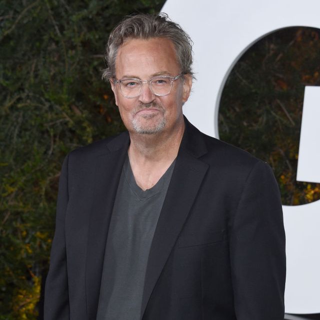 matthew perry smiles at the camera while standing in front of greenery and a large white sign, he wears a black suit jacket over a gray t shirt and clear plastic rimmed glasses