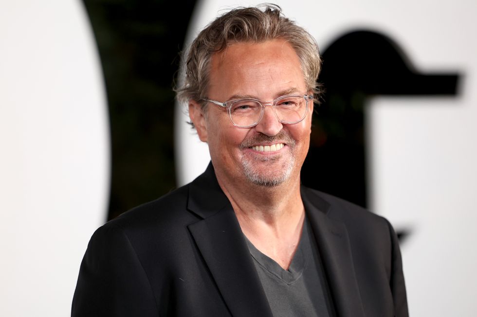 matthew perry smiles and looks right of the camera, he wears a black suit jacket over a gray t shirt and clear plastic rimmed glasses