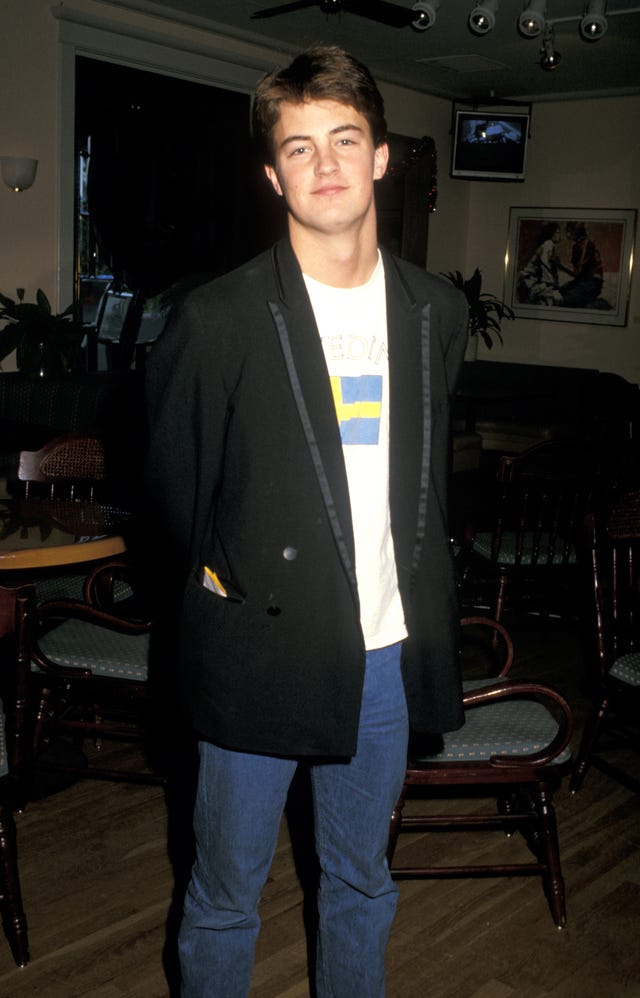 matthew perry stands in a room and looks at the camera with a small smile, he wears a black blazer, a tshirt and jeans