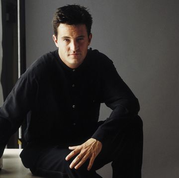 matthew perry sits with one elbow resting on his raised knee, he wears a black long sleeve shirt and pants and looks straight at the camera