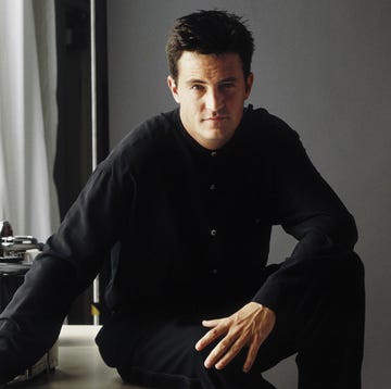 matthew perry sits with one elbow resting on his raised knee, he wears a black long sleeve shirt and pants and looks straight at the camera