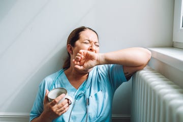 mature multiracial female nurse in blue medical uniform sit on a floor with cup of coffee and yawning covering mouth with hand