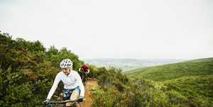 mature woman climbing hill while riding mountain bike on trail with husband