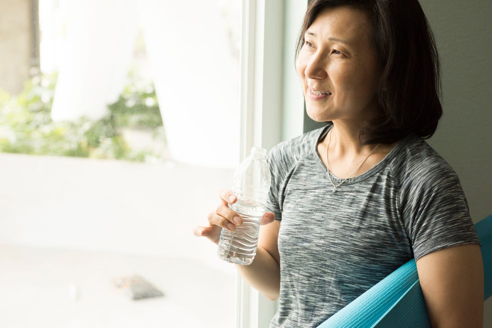 how to look younger mature woman holding rolled up yoga mat and bottle of water looking out of window smiling