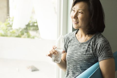 how to look younger mature woman holding rolled up yoga mat and bottle of water looking out of window smiling