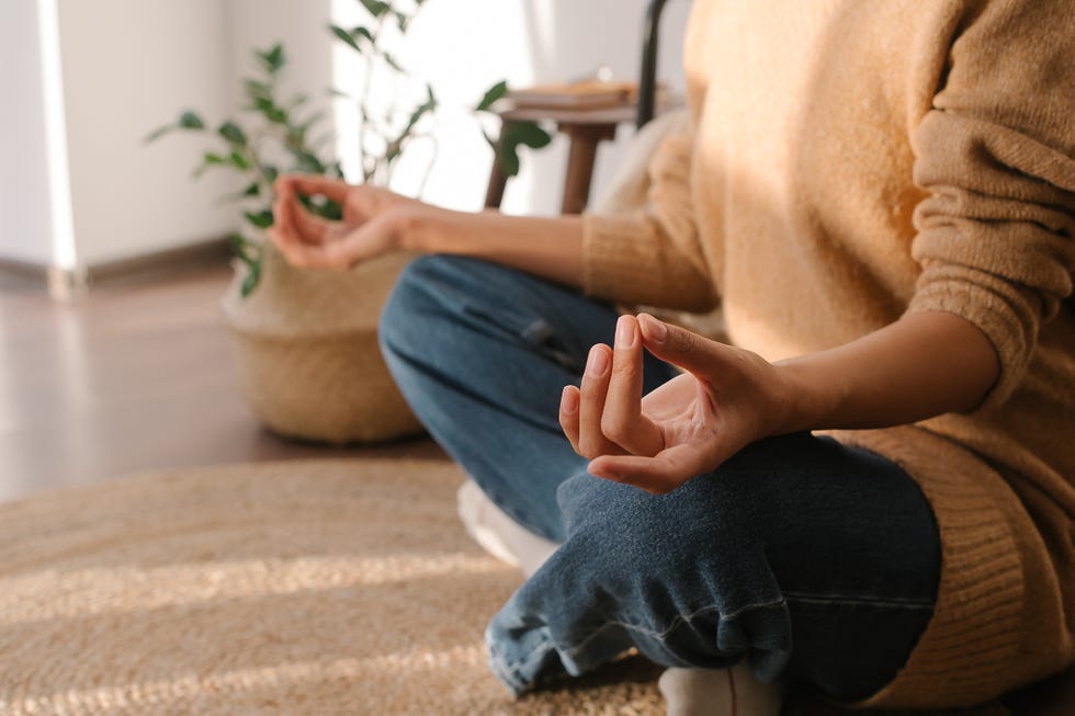 peaceful asian woman meditating while sitting in lotus pose on wicker rug in cozy light bedroom at home, feeling relaxed and tranquil during daily meditation practice in morning
