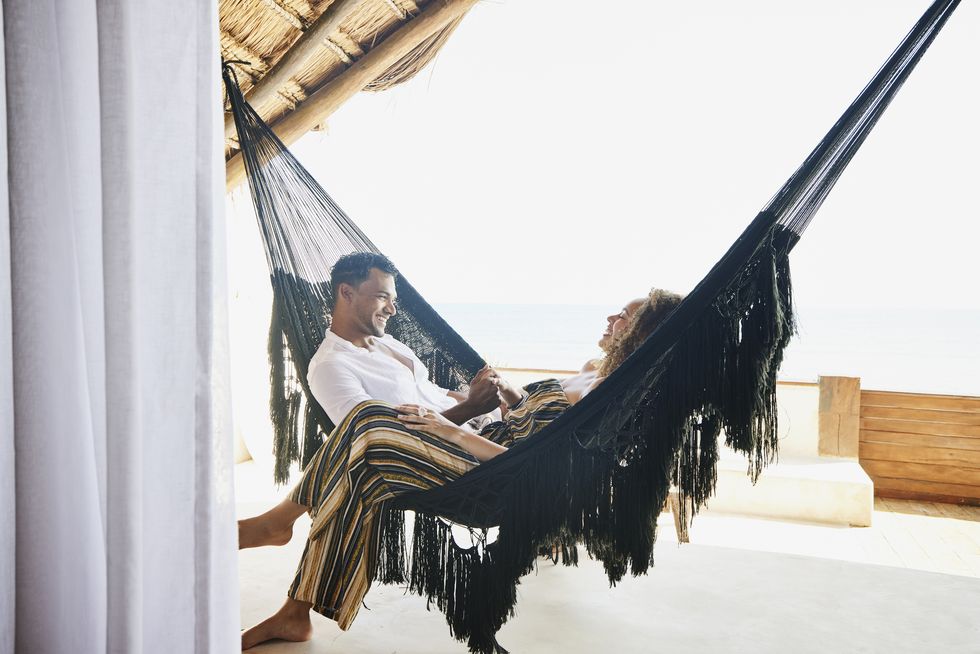 medium wide shot of smiling couple holding hands while sitting in hammock on deck of luxury tropical villa