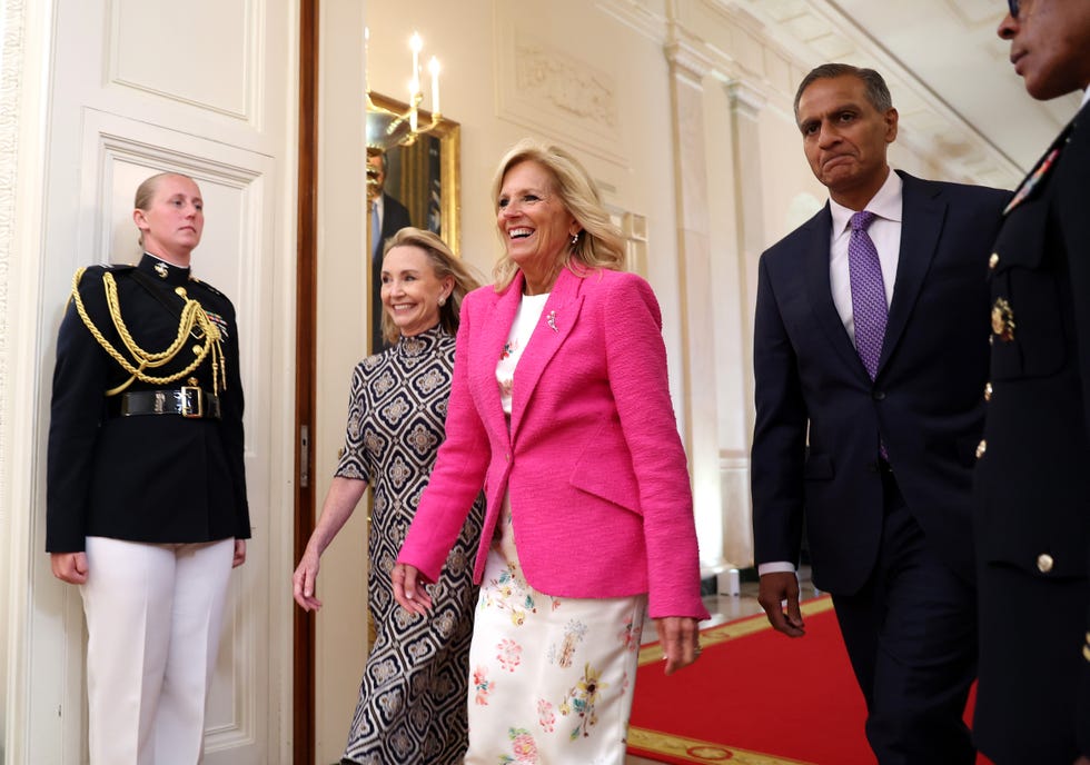 first lady jill biden wearing a pink blazer, walking while smiling next to megan beyer inside the white house