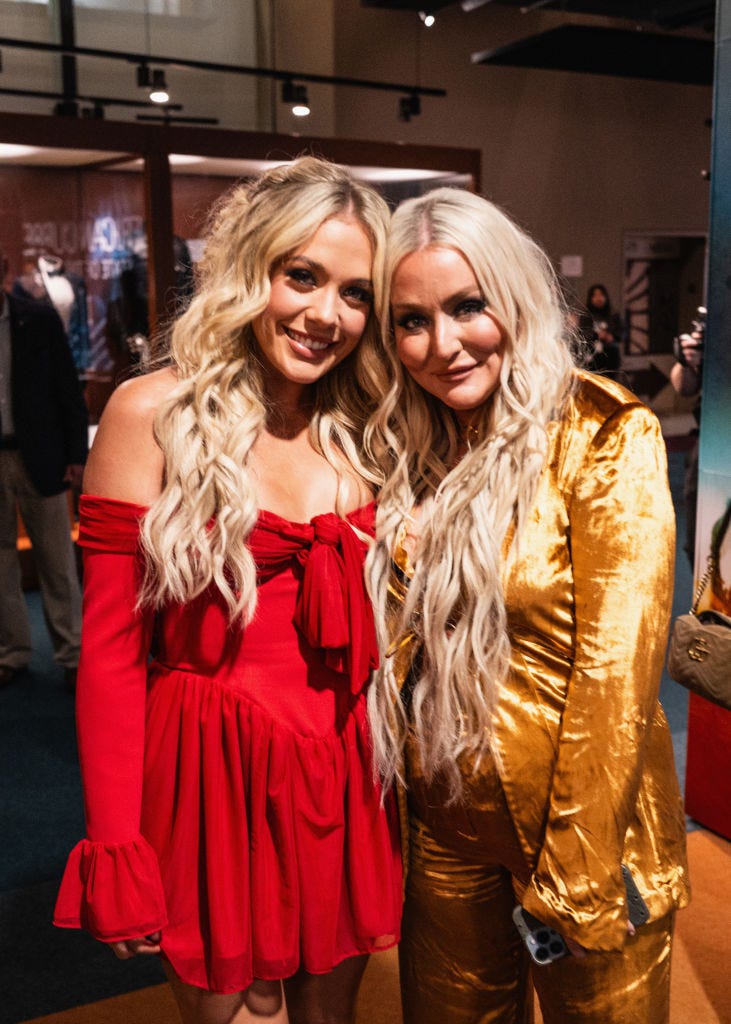 ﻿megan moroney and jessie jo dillon stand together and smile for a photo, moroney wears a red dress, dillon wears a gold velvet outfit