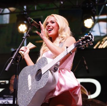 megan moroney smiles as she stand behind a microphone on a stand and holds a trophy in one hand, a bejeweled silver guitar hangs around her shoulders and she wears a pink outfit