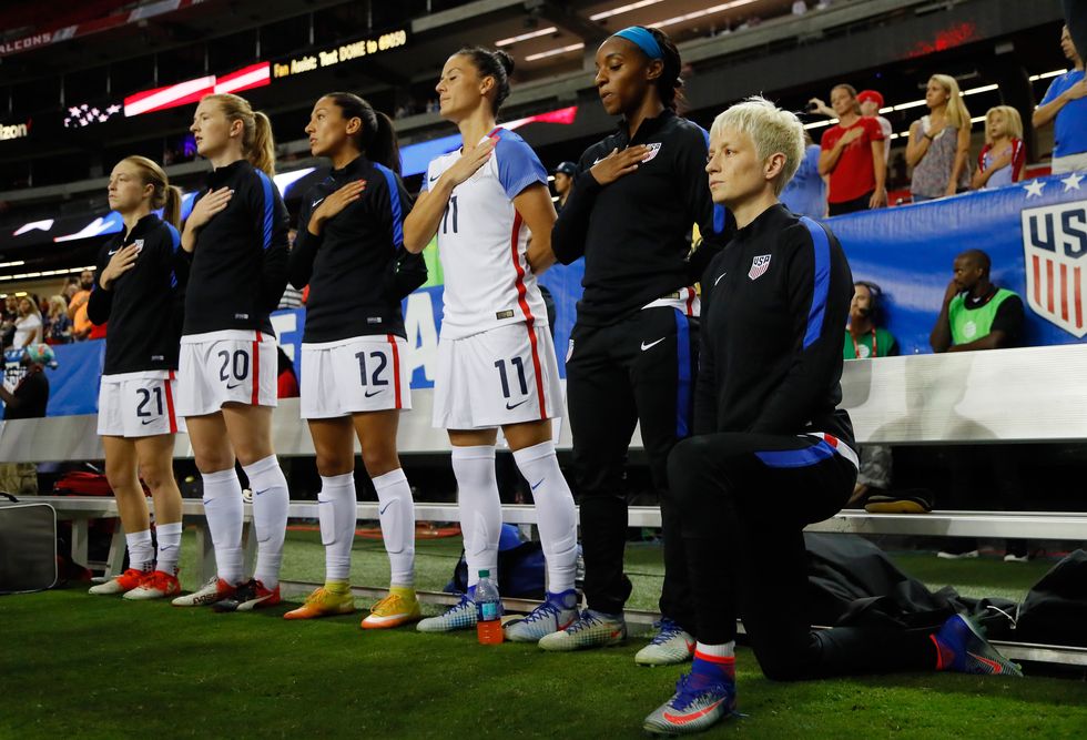 megan rapinoe on one knee for the national anthem while her teammates stand along the bench