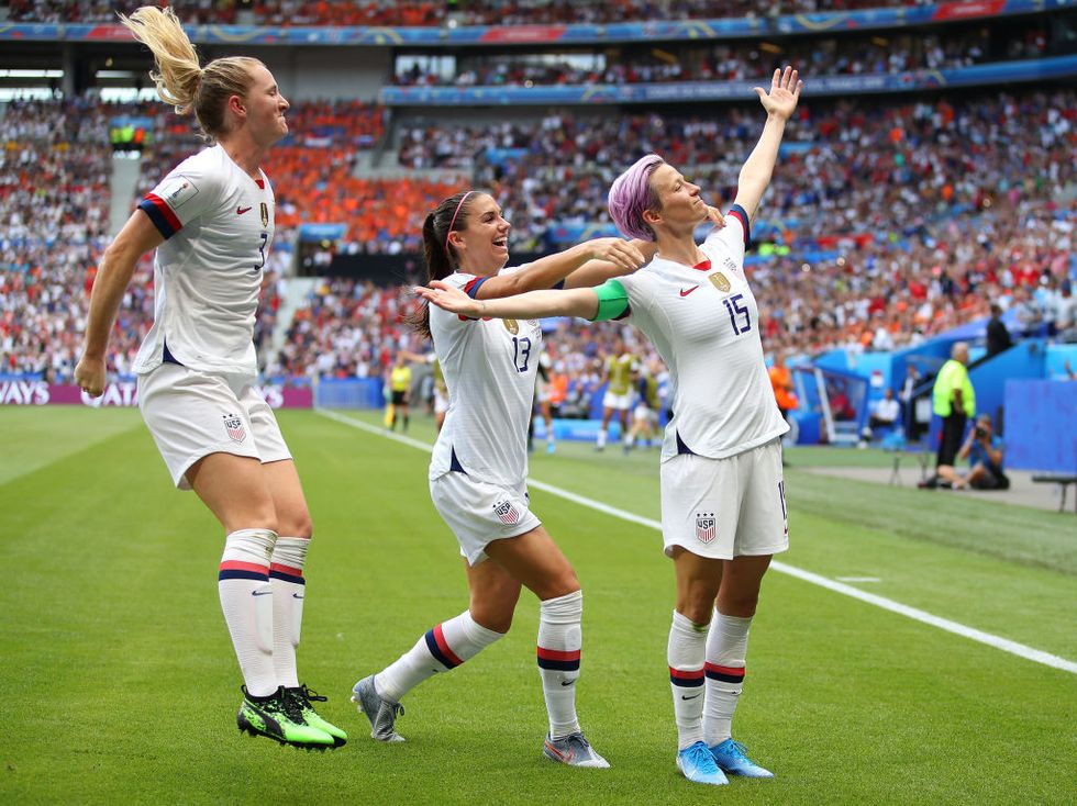 samatha mewis and alex morgan run to megan rapinoe who stands on a soccer field is her arms extended and head looking up, all three wear white usa team uniforms and soccer cleats