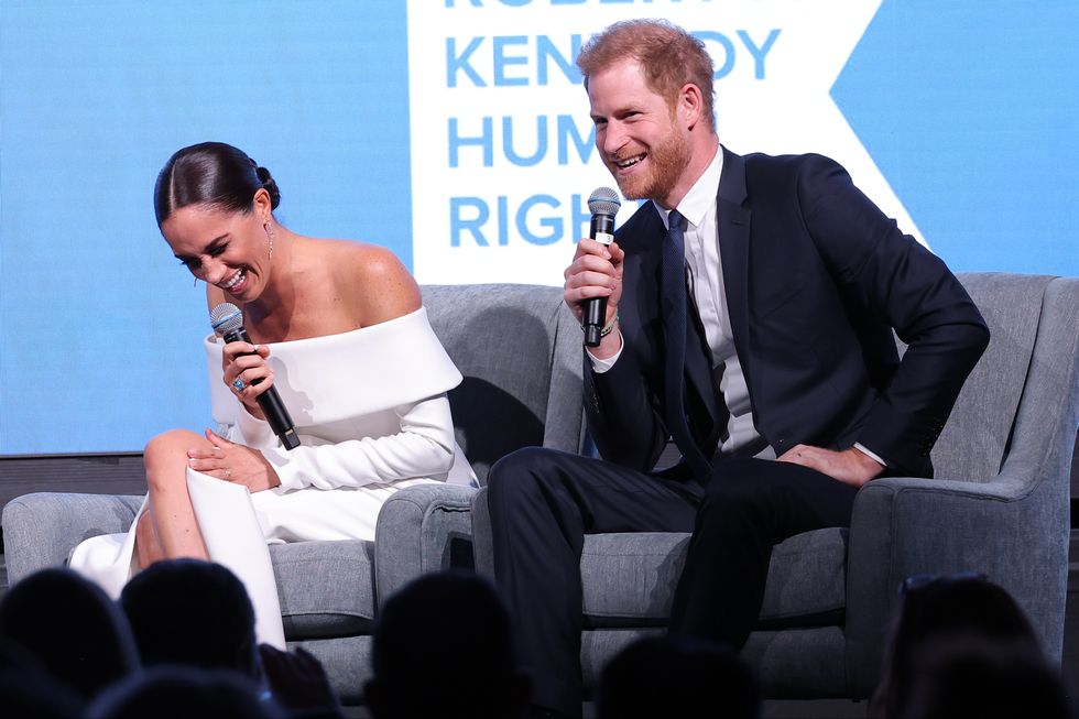meghan markle sitting in a chair laughing as prince harry speaks to an audience