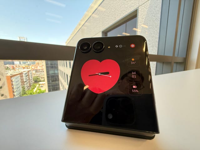 un teléfono móvil motorola razr 60 ultra plegable con un corazón en la pantalla externa sobre una mesa al lado de una ventana con edificios al fondo