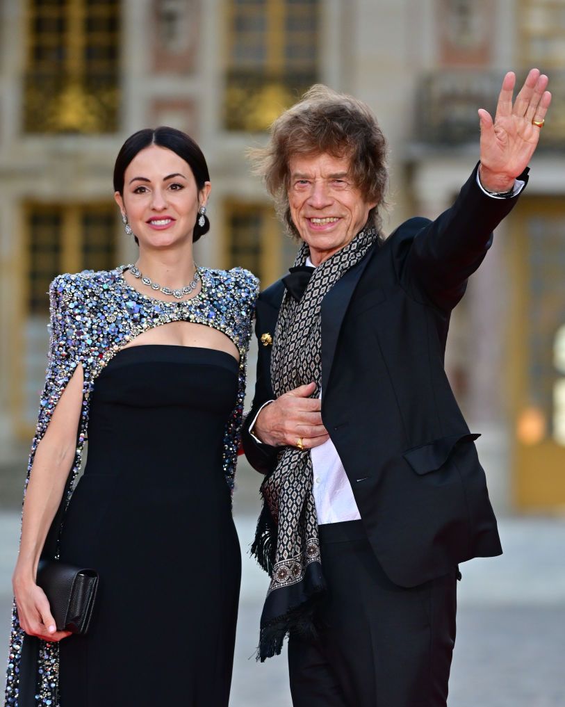 melanie hamerick and mick jagger smile and look past the camera while standing next to each other outside, she wears a bejeweled cape, strapless black dress, and jewelry and holds a black clutch purse, he wears a dark suit with a patterned scarf and waves one hand