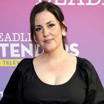 melanie lynskey, a woman stands looking at the camera with a neutral facial expression, she has dark hair worn back in a ponytail and wears a black dress