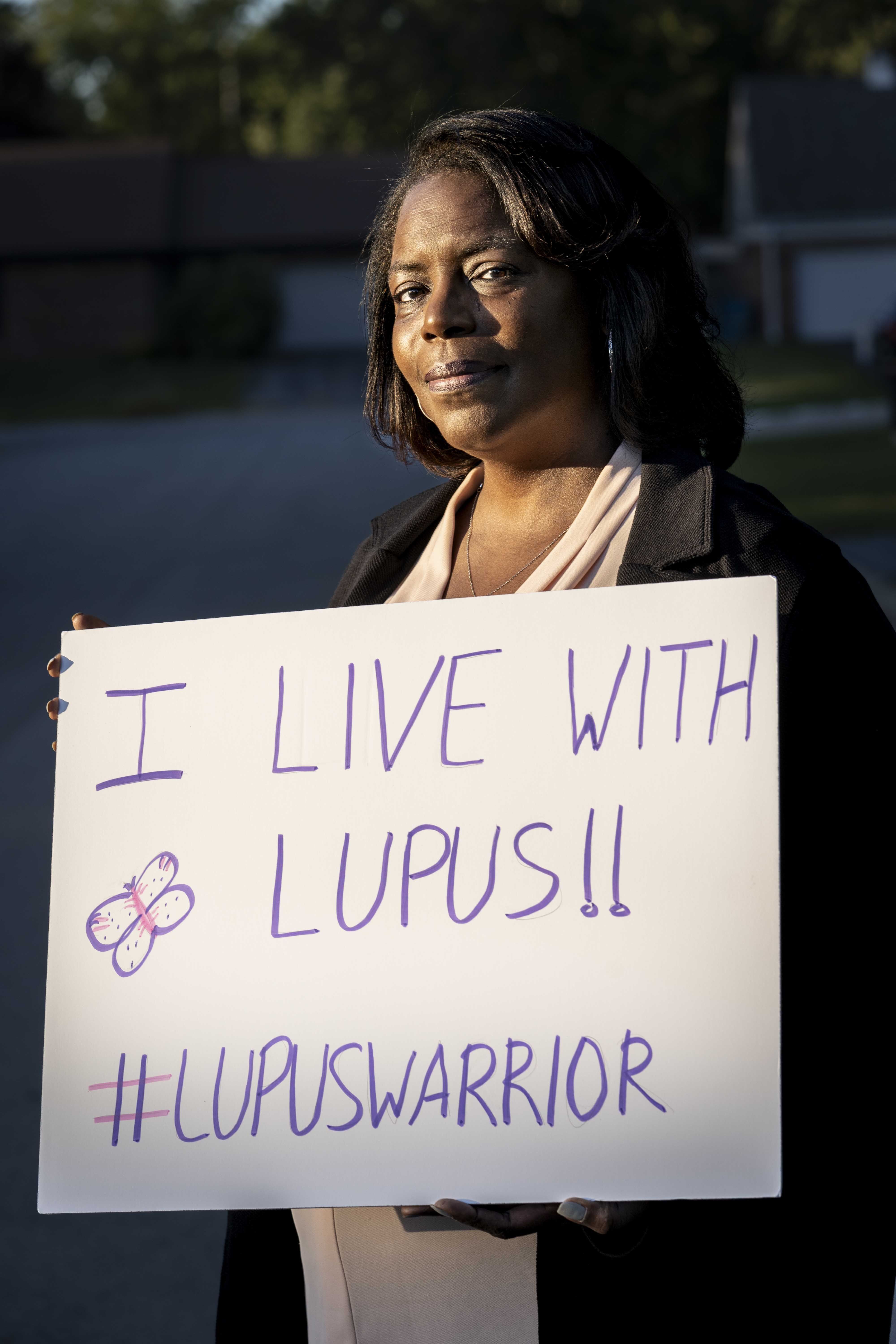 melanie hall at her home in crete, illinois, holding a white sign that has the hashtag lupuswarrior