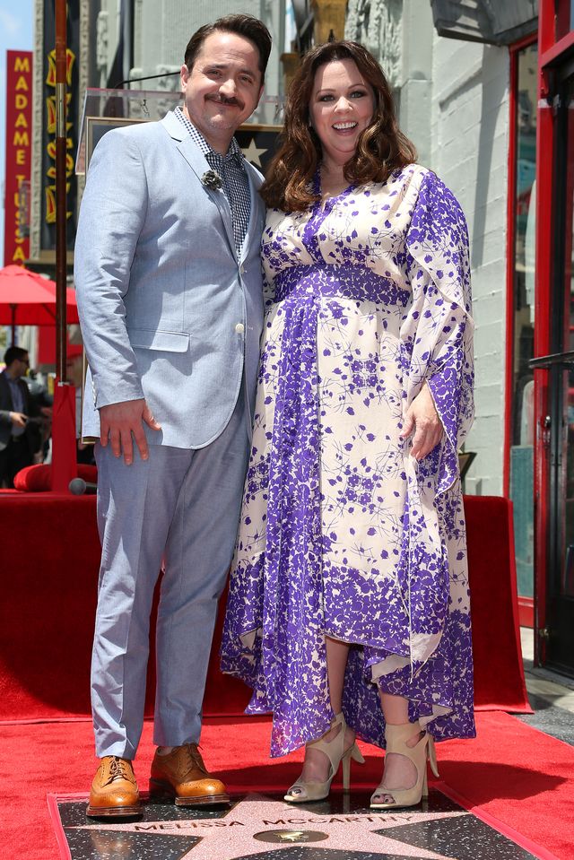 ben falcone and melissa mcccarthy smile as they stand behind a hollywood star bearing mccarthys name, he wears a light blue suit with a blue checkered shirt, she wears a white and purple floral dress
